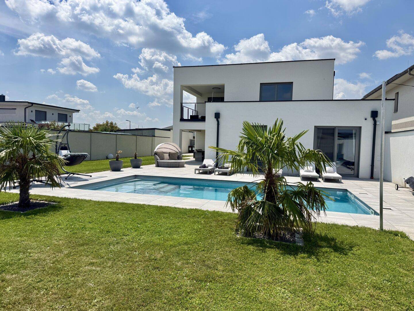 Gartenbereich mit Pool, Palmen und moderner Villa unter strahlend blauem Himmel.