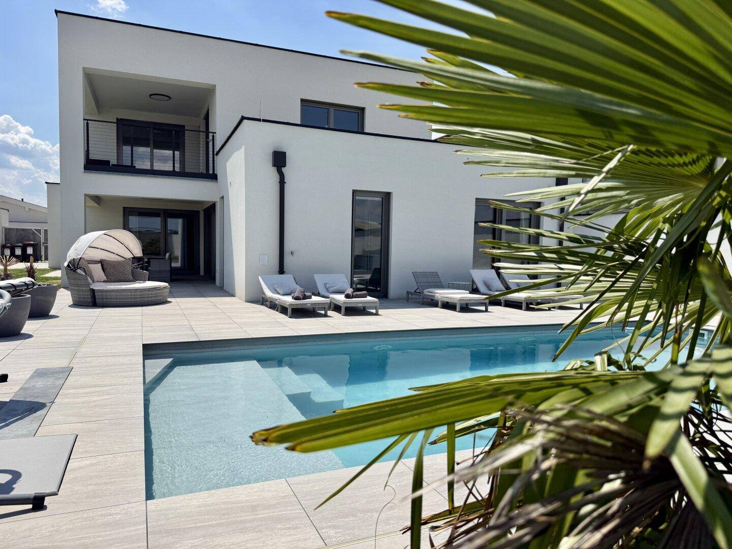 Rückansicht eines modernen Hauses mit Pool, Terrasse und Palmen unter blauem Himmel.