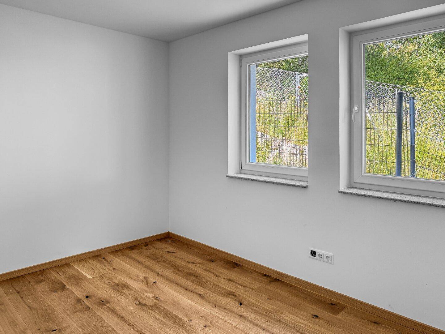 Geräumiges Schlafzimmer mit Holzboden und zwei Fenstern mit Blick ins Grüne.