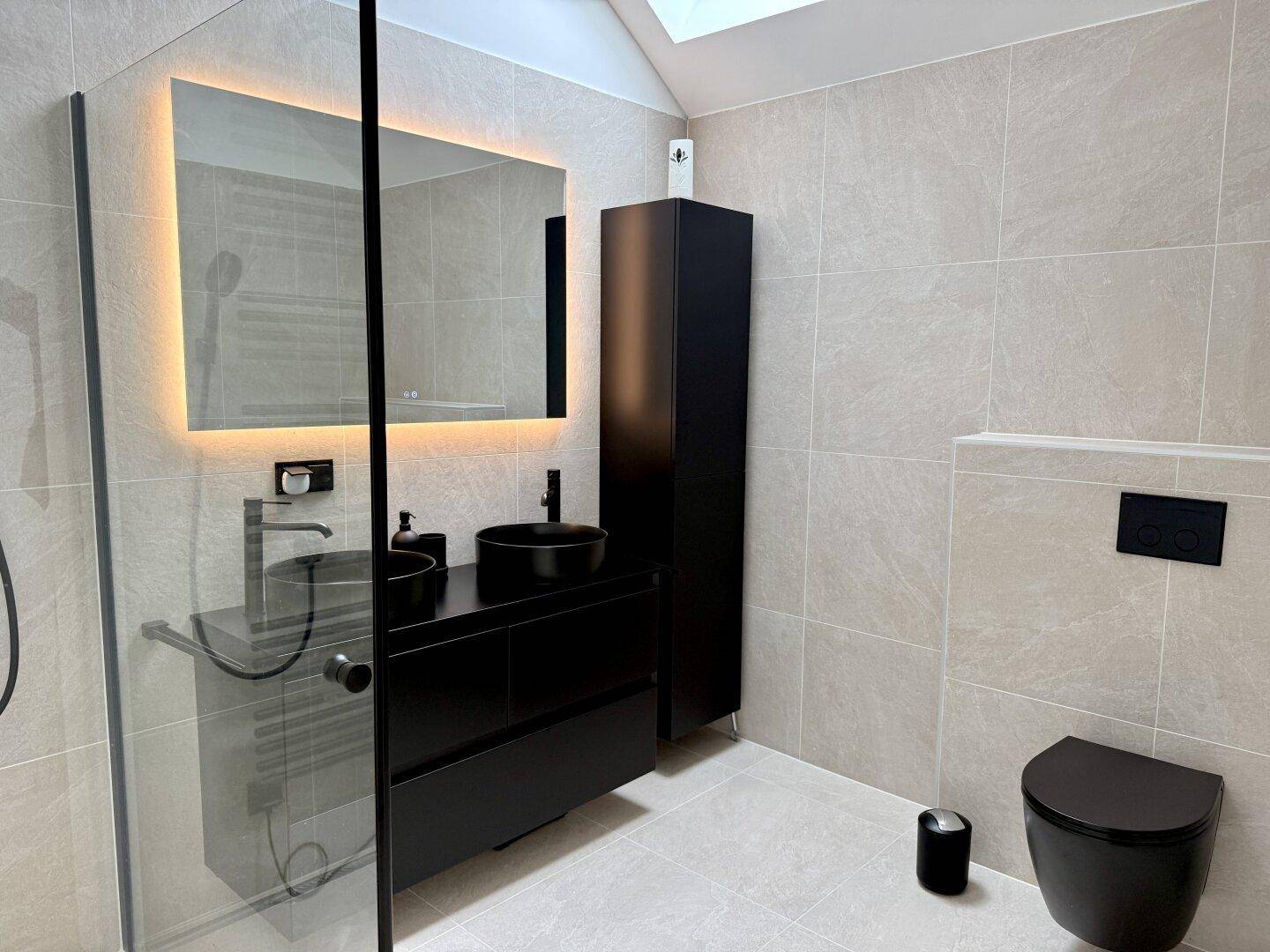 Modernes Badezimmer mit schwarzem Waschbecken, Toilette, Dusche und beleuchtetem Spiegel.