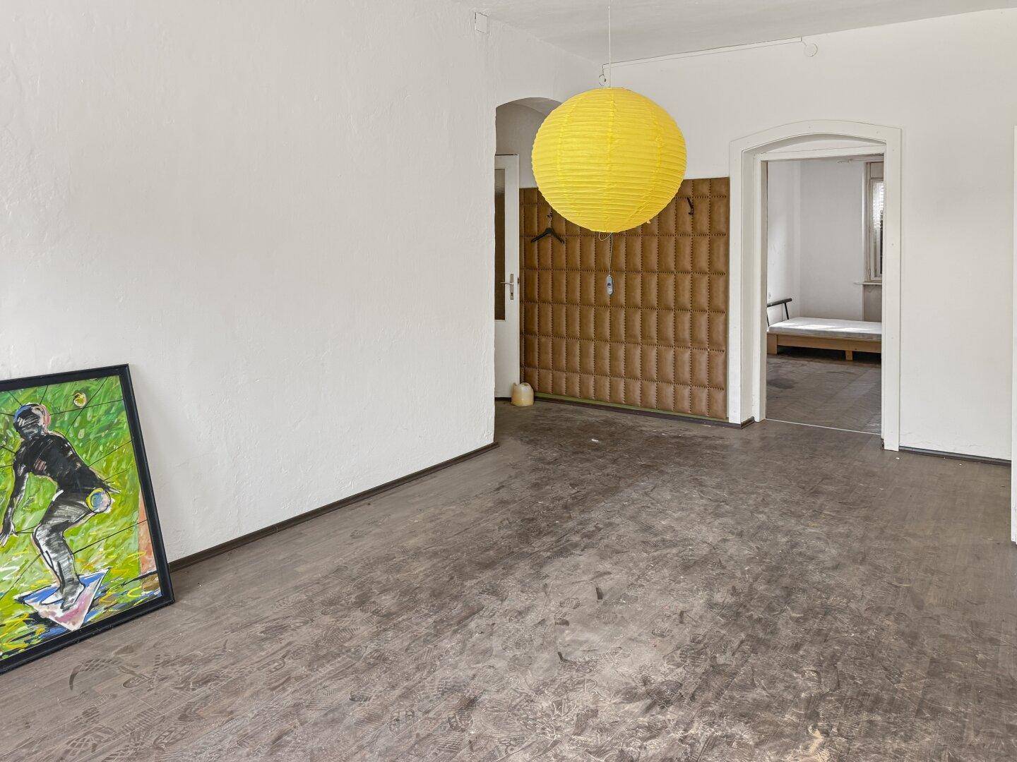 Leerer Raum mit Laminatboden, weißen Wänden und einer gelben Deckenlampe, offen zu einem weiteren Zimmer.