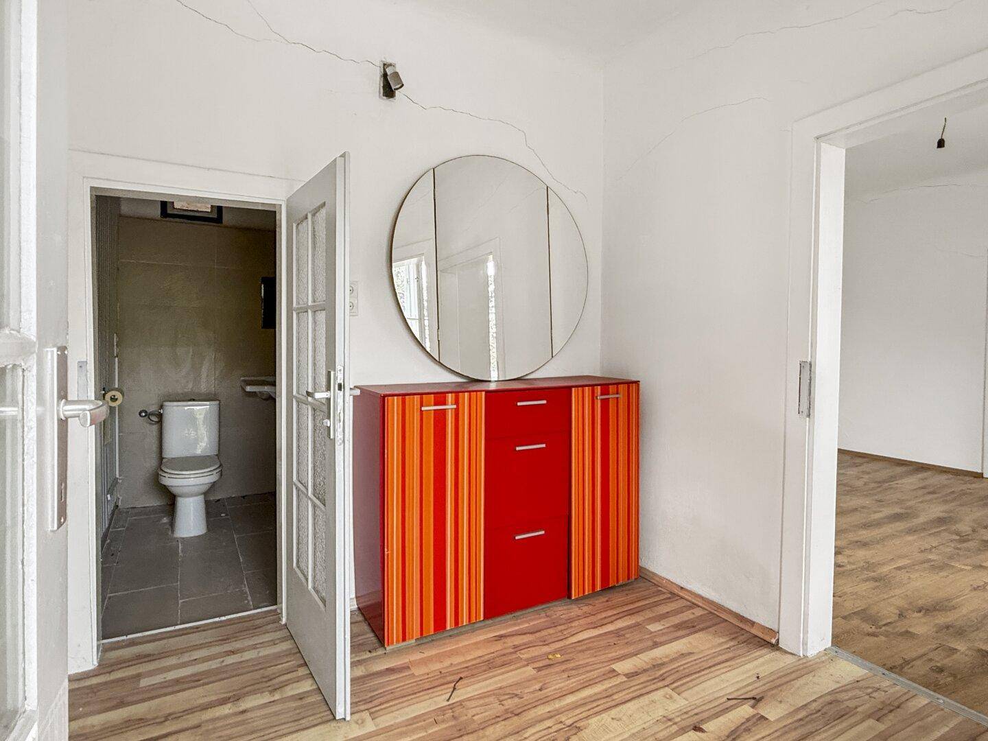 Flur mit Laminatboden, einem farbenfrohen Schrank, rundem Spiegel und Zugang zu einem Badezimmer.