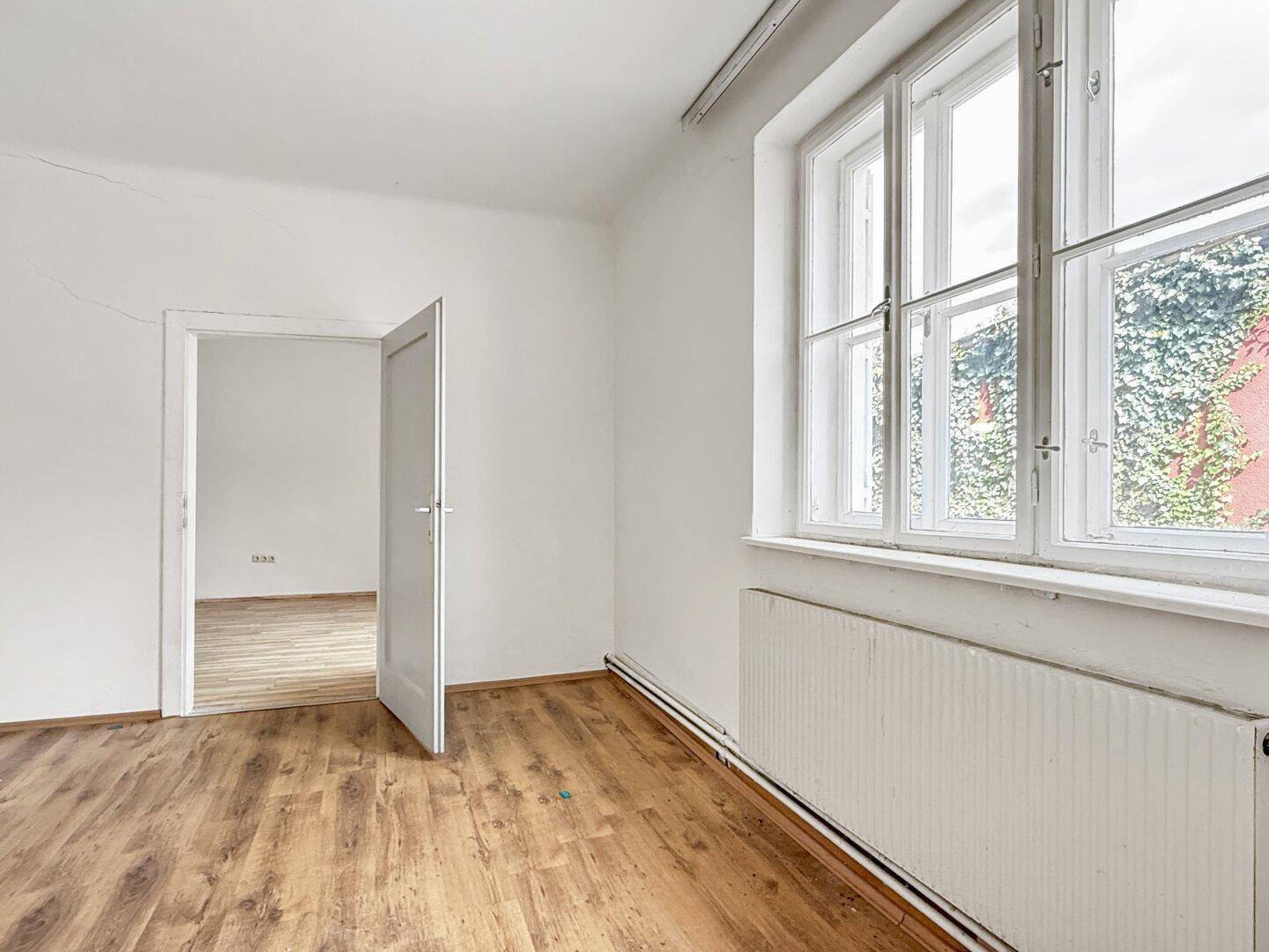 Lichtdurchflutetes Zimmer mit Laminatboden, Heizkörper und Blick ins Grüne durch die Fenster.