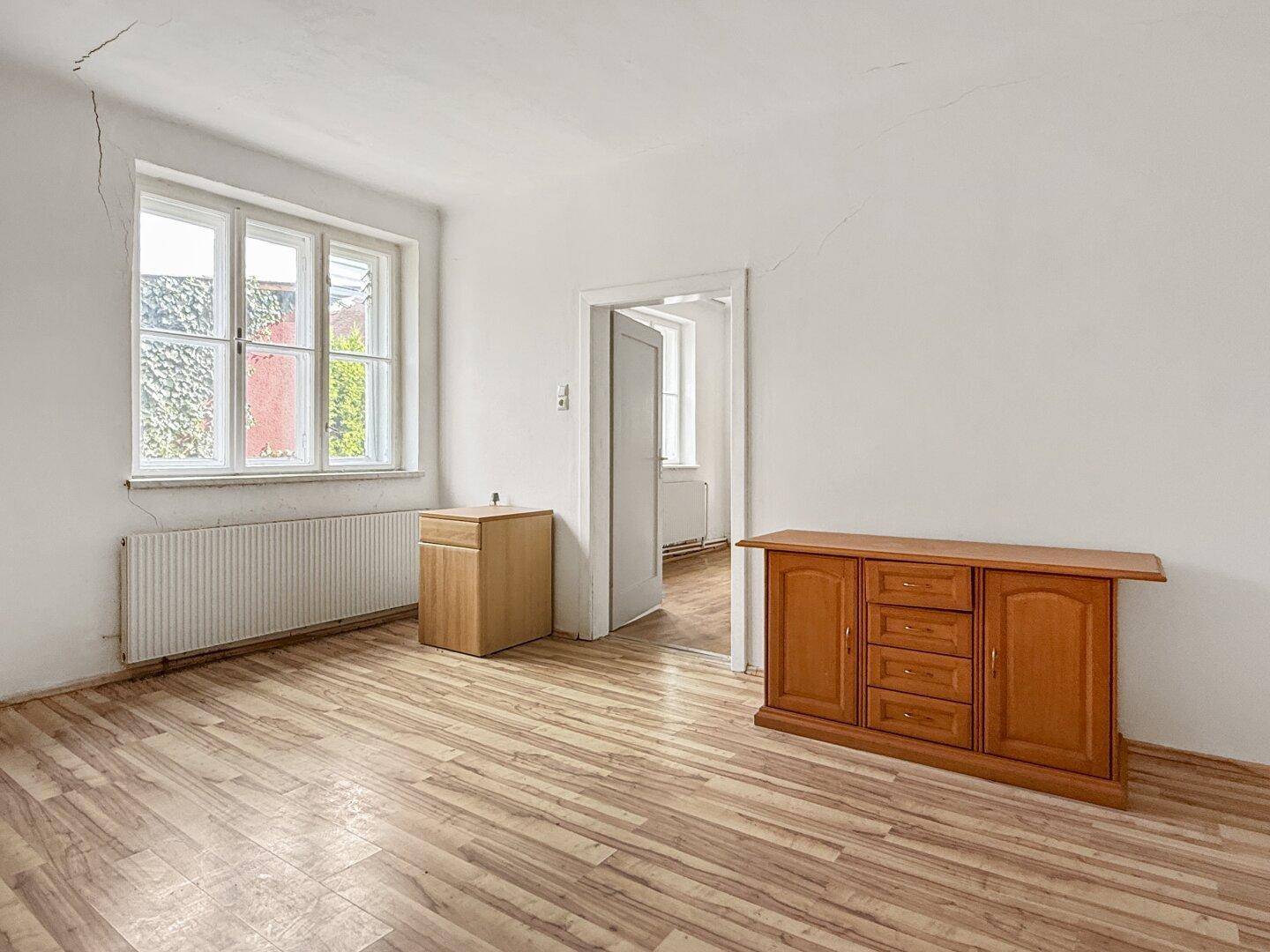 Helles Zimmer mit Laminatboden, Fenster und zwei Holzmöbelstücken, die den Raum schmücken.