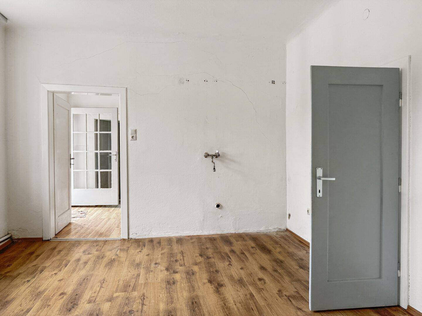 Leerer Raum mit Laminatboden, vorbereiteten Anschlüssen für eine Küche und Zugang zu einem weiteren Zimmer.