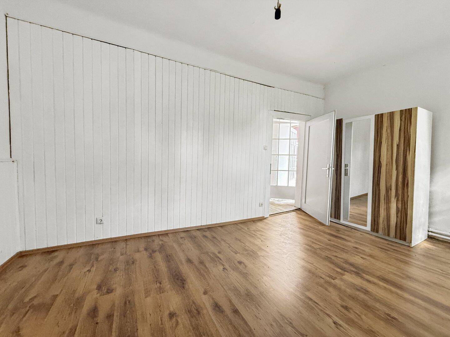Heller Raum mit Laminatboden, weißen Holzpaneelen und einem Kleiderschrank mit Spiegeltür.