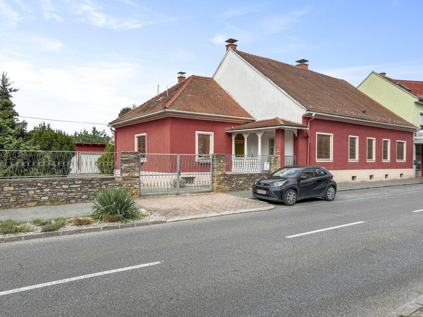 Ansicht des roten Hauses von der Straße mit Vorgarten, Zaun und einem geparkten Auto davor.