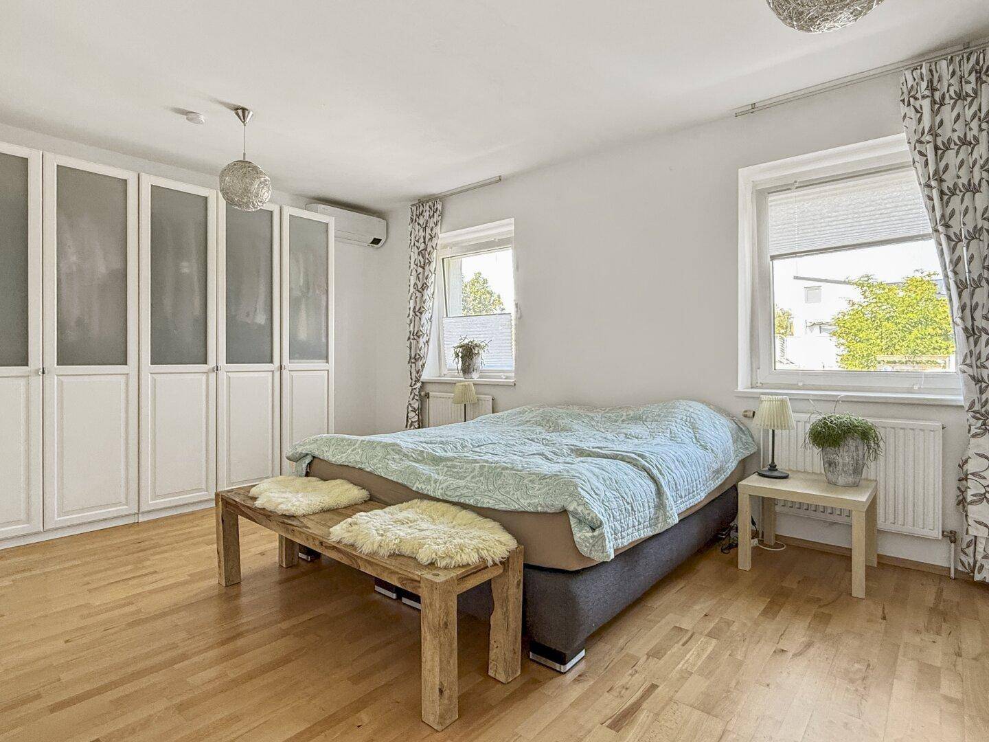 Helles Schlafzimmer mit Doppelbett, Holzboden und großen Fenstern.