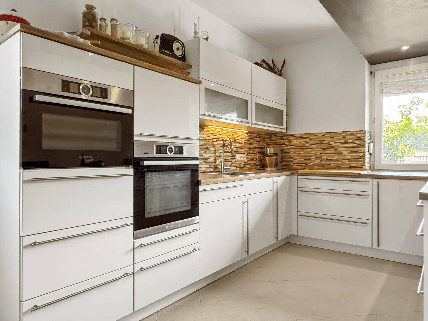 Moderne Einbauküche mit weißen Hochglanzfronten, zwei Öfen und Mosaikfliesen als Spritzschutz.