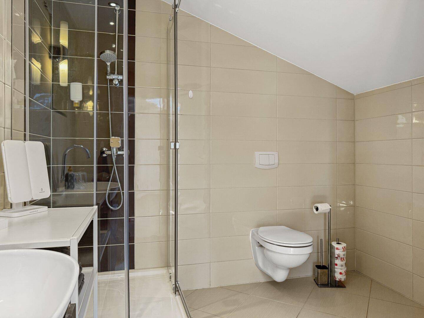 Badezimmer mit beigefarbenen Fliesen, Wand-WC und Duschkabine im Dachgeschoss.