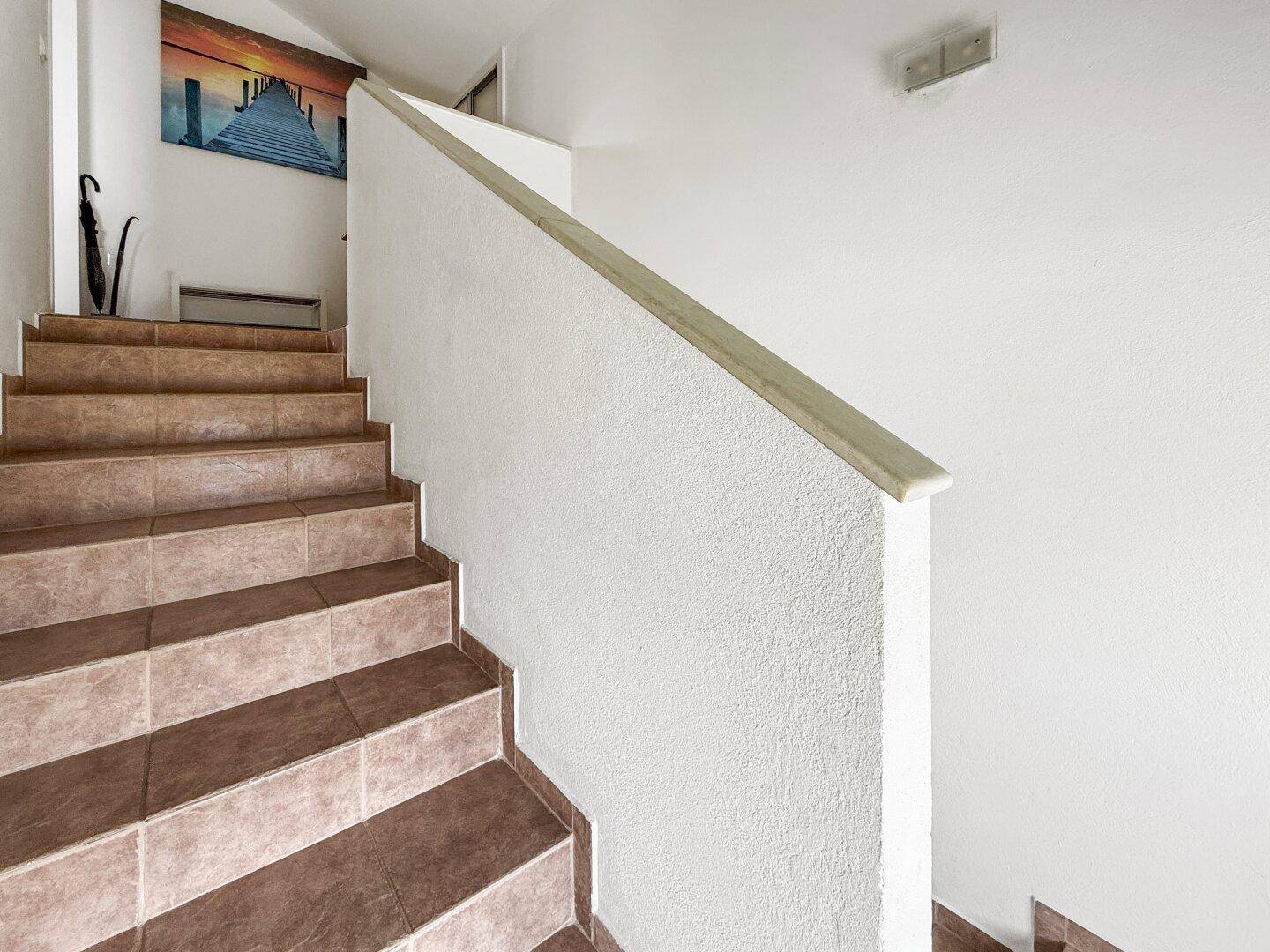 Geflieste Treppe mit hellem Geländer und einem Bild an der Wand, die in die obere Etage führt.