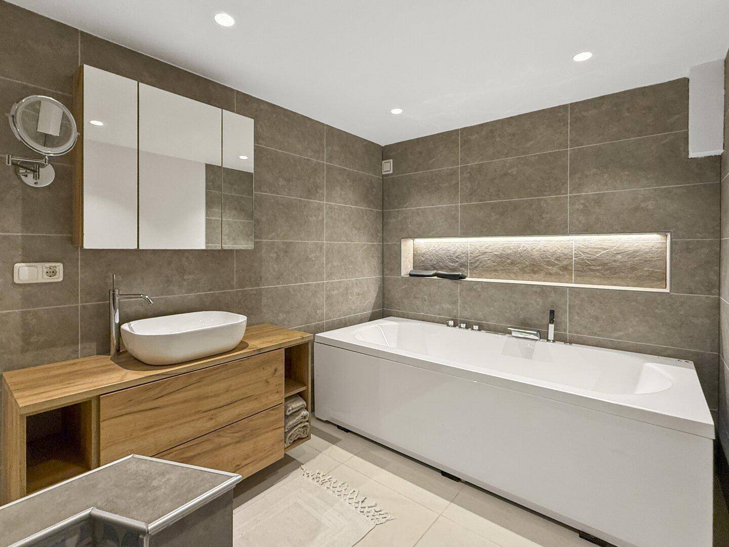 Elegantes Badezimmer mit Badewanne, Holzwaschtisch, Spiegelschrank und indirekter Beleuchtung.