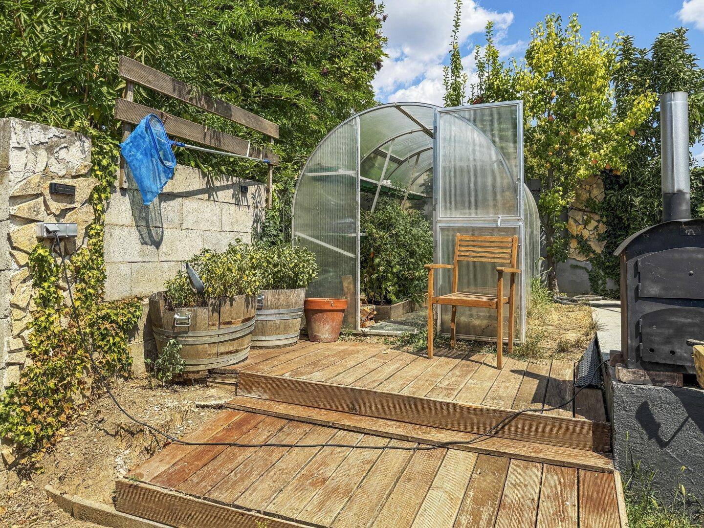 Gartenbereich mit Gewächshaus, Holzdeck und einem Außenofen, ideal für Hobbygärtner und Grillabende.