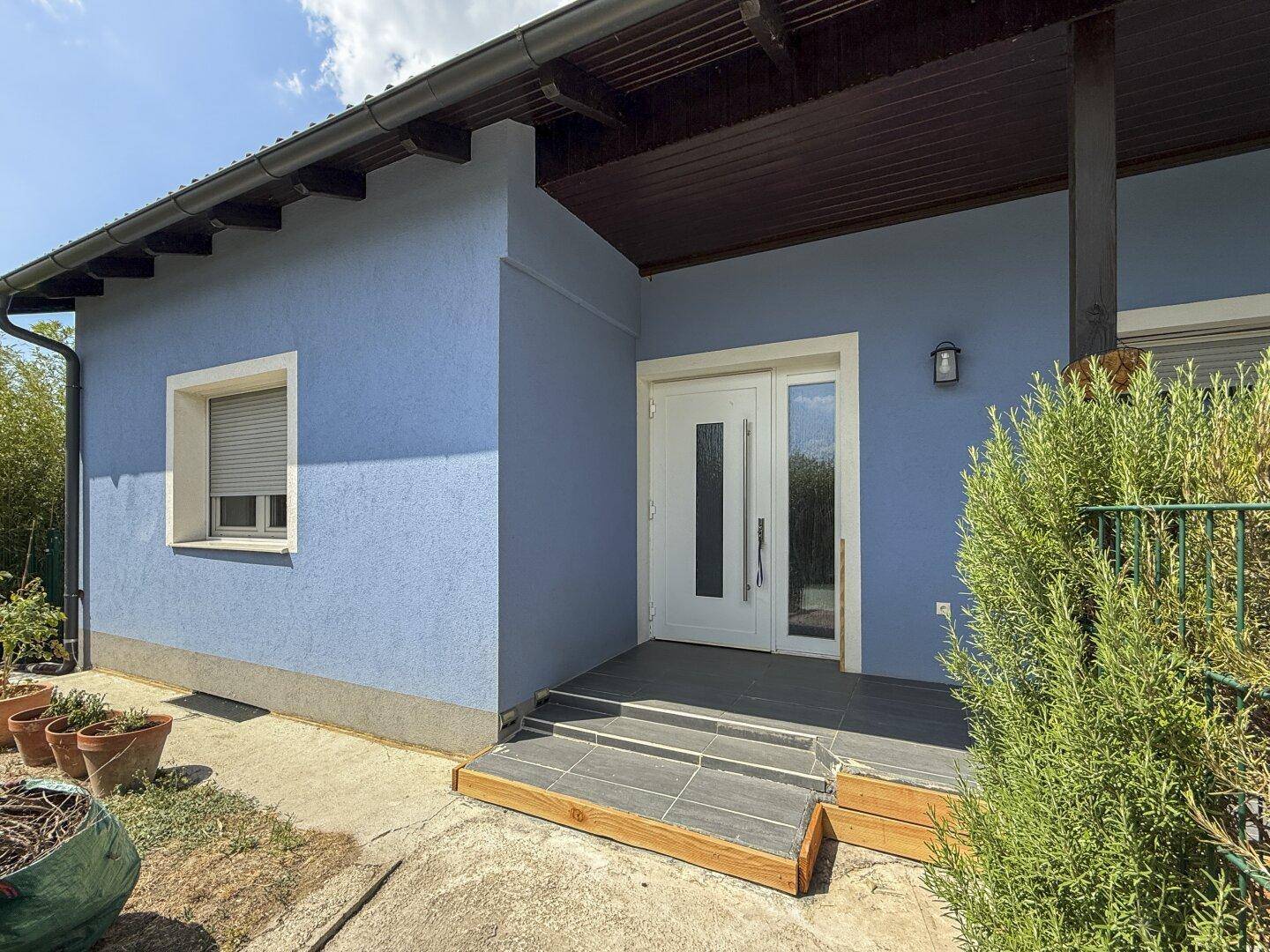 Eingangsbereich des blauen Hauses mit weißer Haustür und kleinen Stufen, umgeben von Grün.