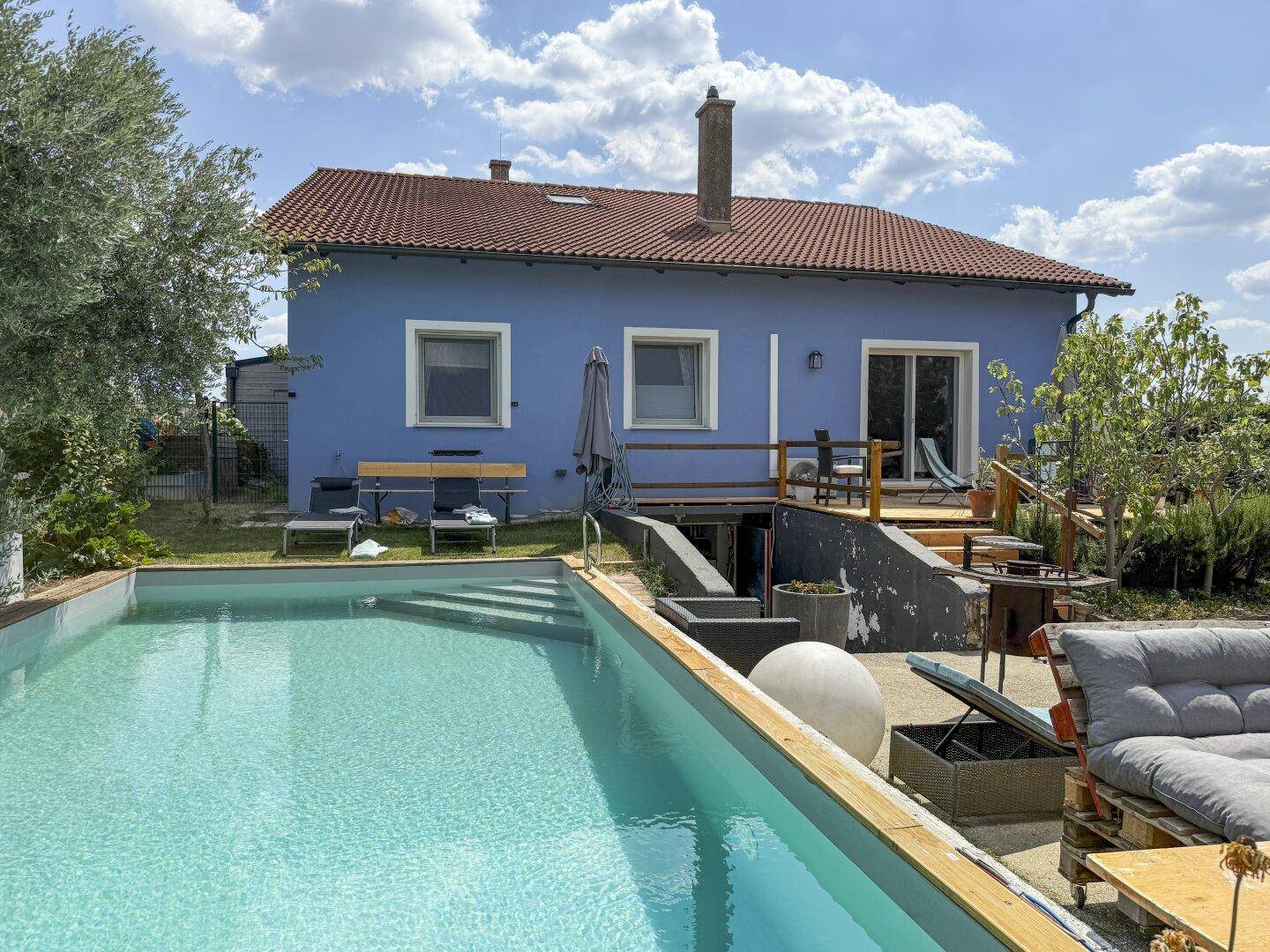 Blaues Haus mit großem Außenpool und Holzterrasse, umgeben von einem gepflegten Garten.