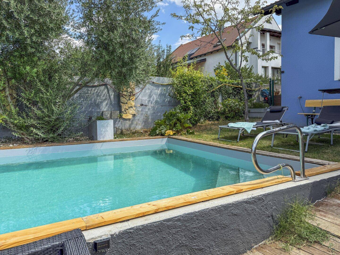 Seitlicher Blick auf den Poolbereich mit Treppeneinstieg und der blauen Hausfassade im Hintergrund.