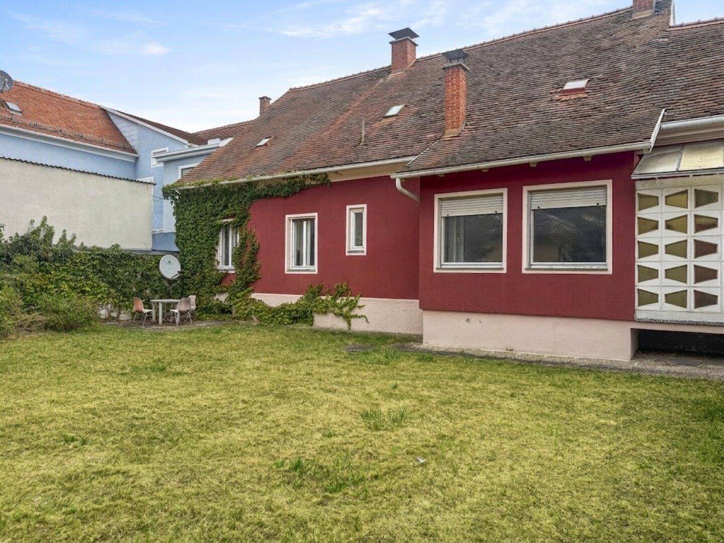 Hinterhof mit Rasenfläche, roter Hausfassade und einer kleinen Sitzecke im Schatten.