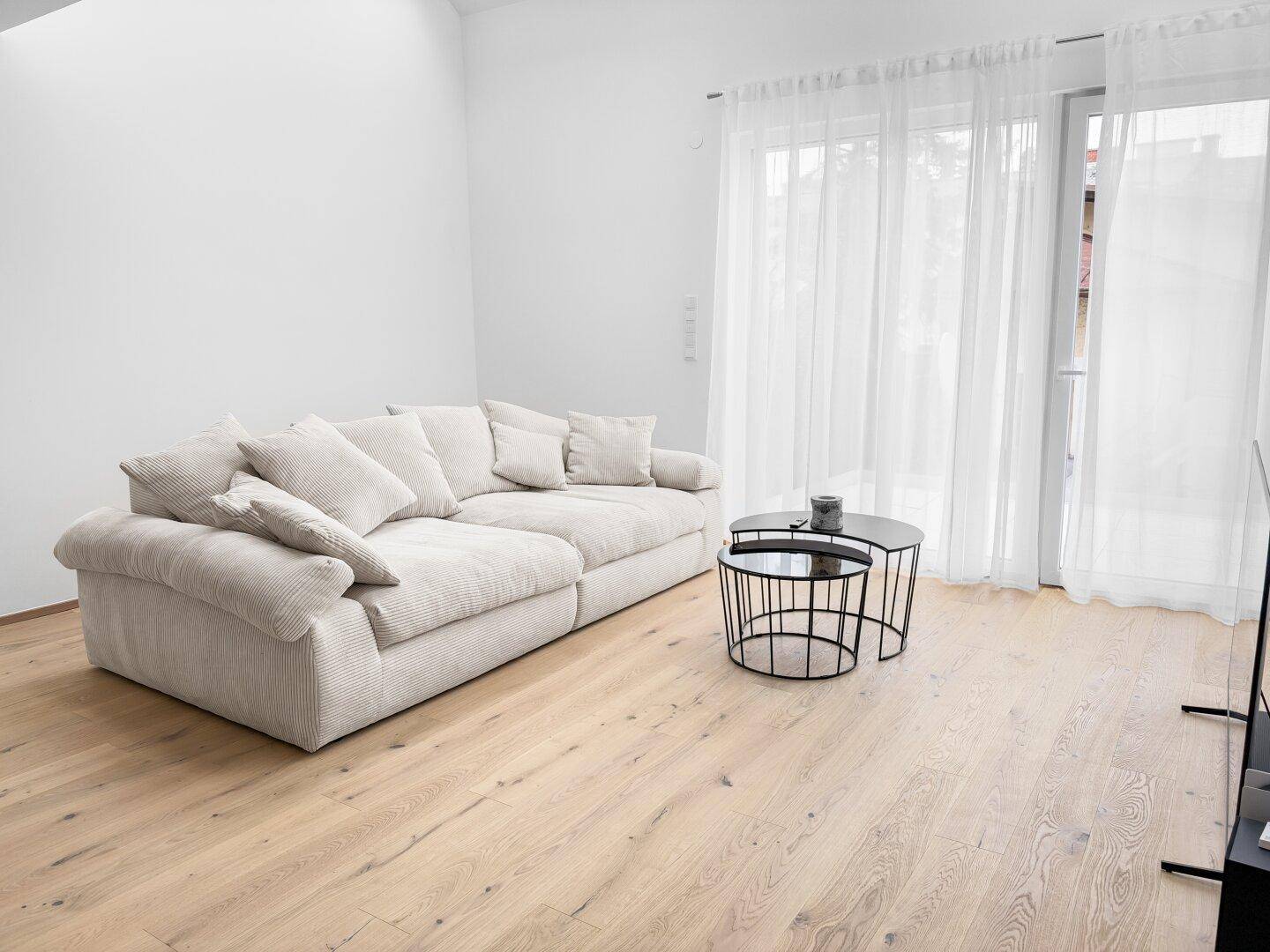 Heller Wohnbereich mit bequemem Sofa, Eichenparkett und großen Fenstern mit transparenten Vorhängen.