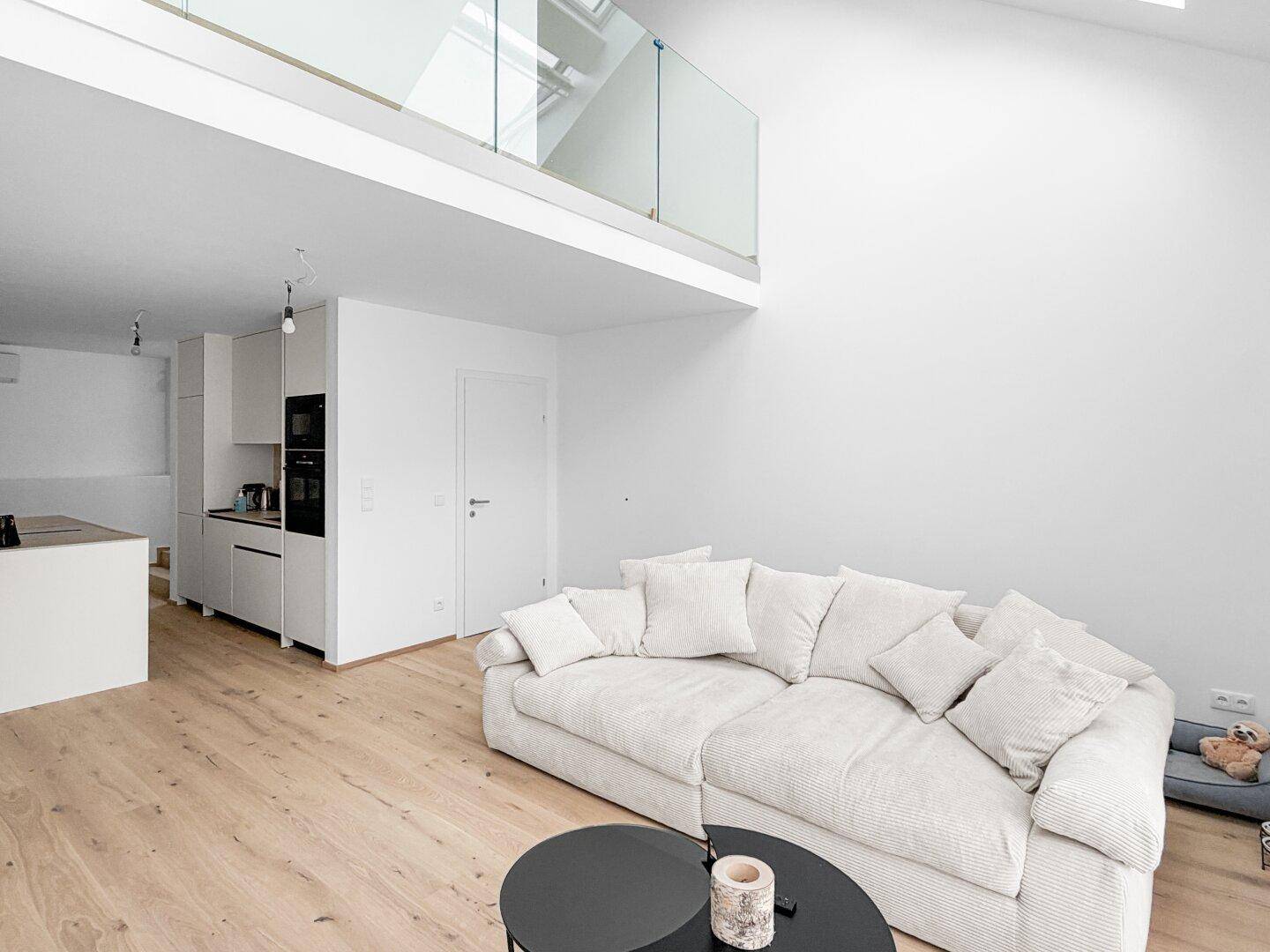 Heller Wohnbereich mit großem Sofa, Holzfußboden und offener Galerie mit Glasgeländer.