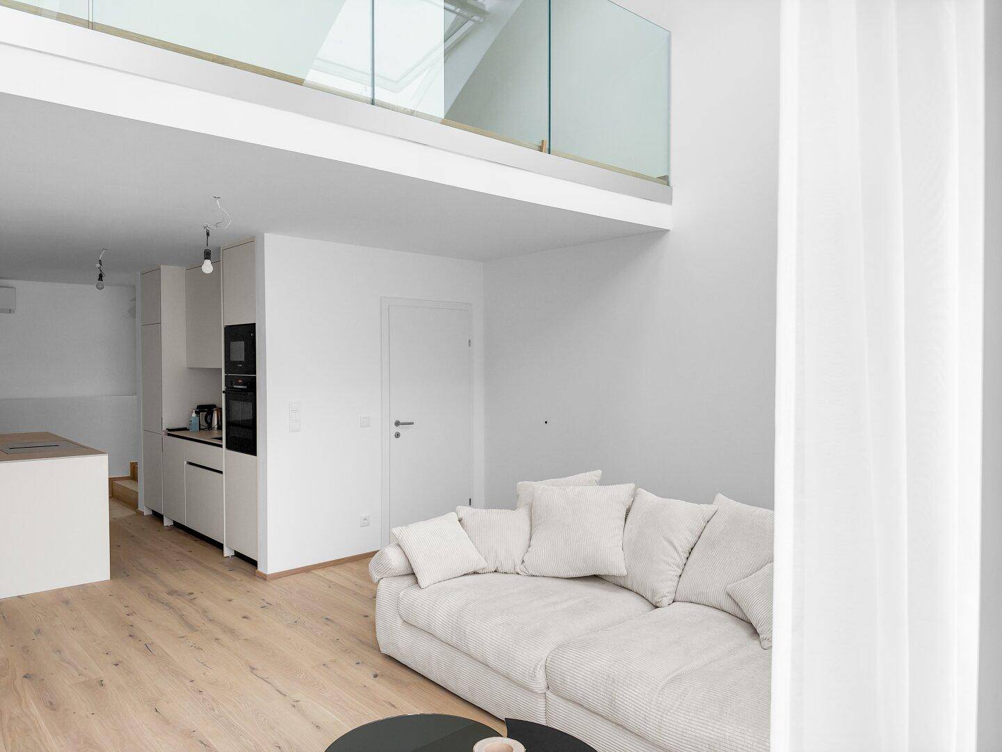 Geräumiger Wohnbereich mit beigem Sofa, Holzparkett und offener Galerie mit elegantem Glasgeländer.