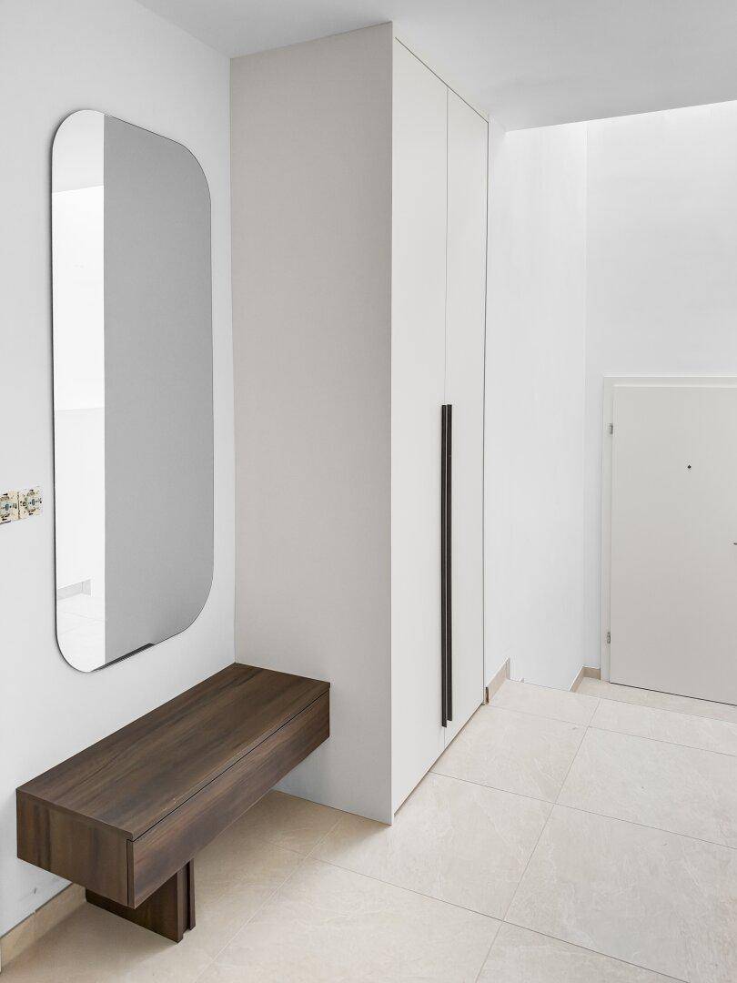 Minimalistischer Flur mit weißer Garderobe, großem Spiegel und schwebender Holzbank am Eingang.