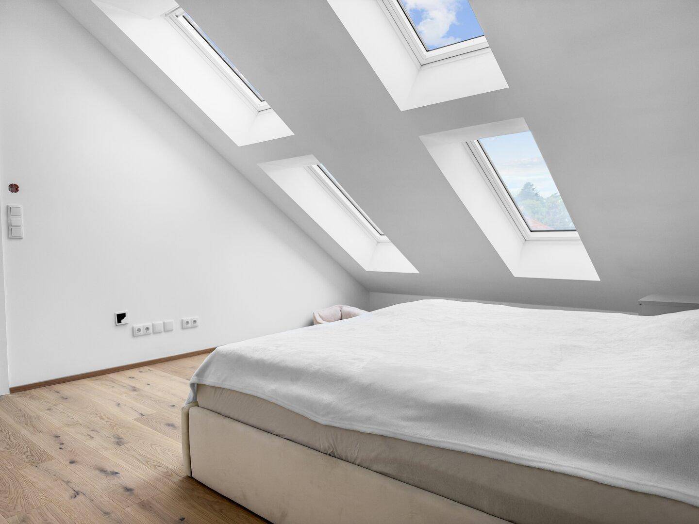 Schlafzimmer unter der Dachschräge mit mehreren Dachfenstern und hellem Holzfußboden.