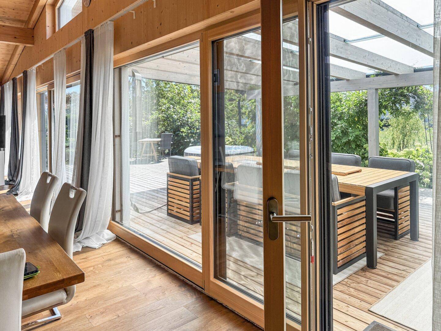Essbereich mit großen Glasfenstern, die den Zugang zur überdachten Holzterrasse ermöglichen und viel Licht spenden.