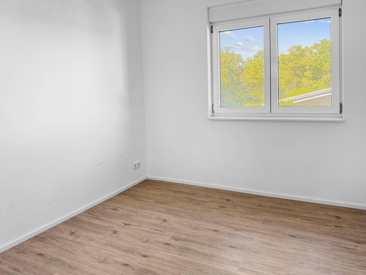 Helles Zimmer mit Holzboden und einem Fenster, das einen Blick ins Grüne bietet.