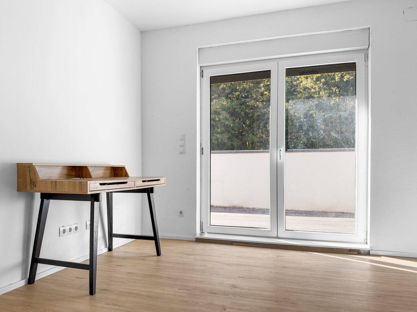 Heller Raum mit Holzboden und großem Fenster, das viel Tageslicht hereinlässt, mit Schreibtisch.