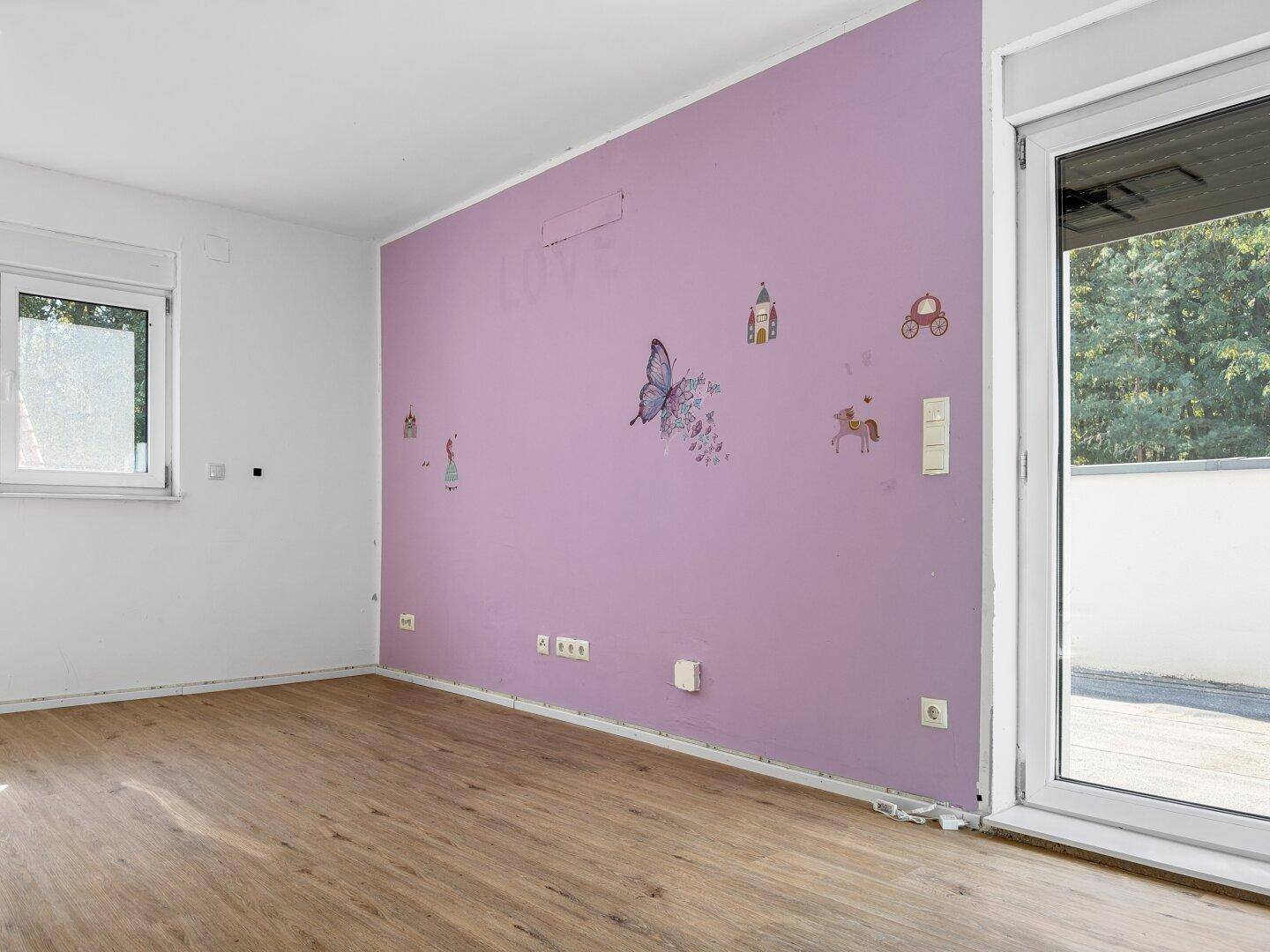Kinderzimmer mit lila Akzentwand und verspielten Wandstickern, hell durch große Fenster.