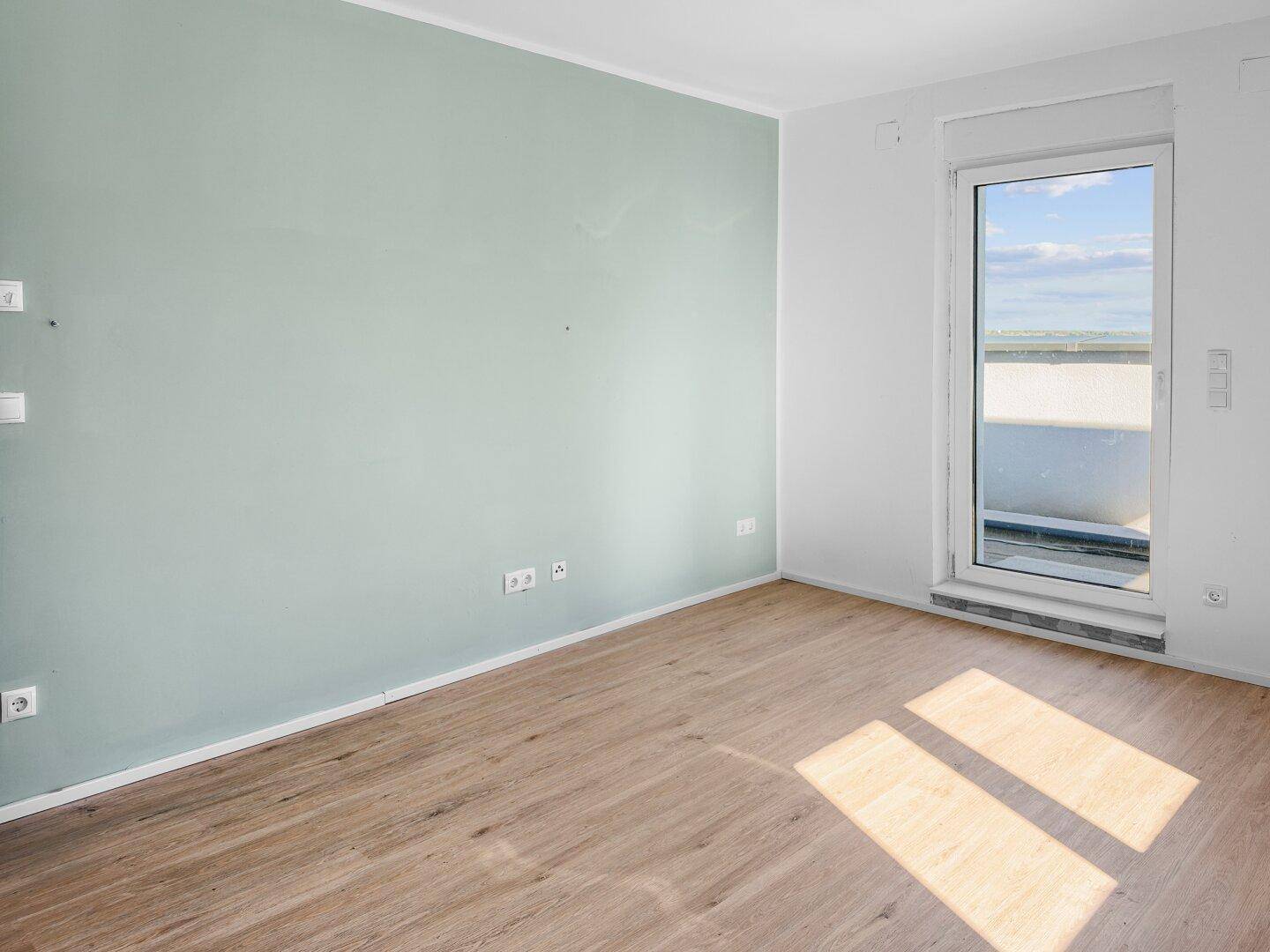 Helles Zimmer mit hellgrüner Akzentwand und Holzboden, mit Zugang zu einem Außenbereich.