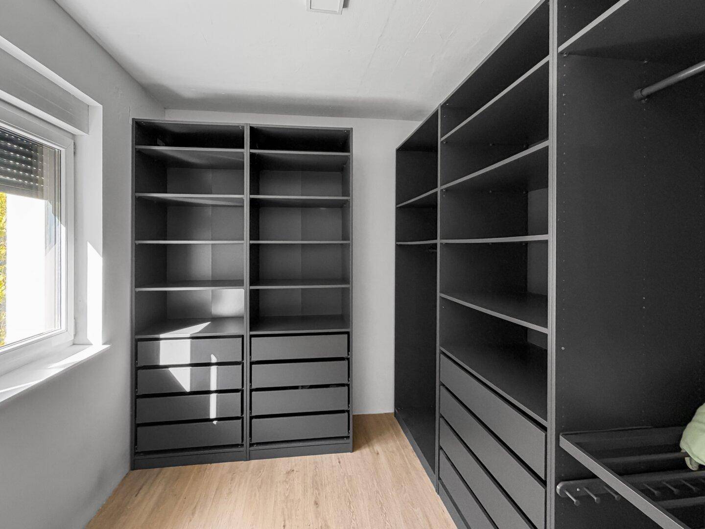 Geräumiger begehbarer Kleiderschrank mit modernen dunklen Regalen und Schubladen, hell durch Fenster.