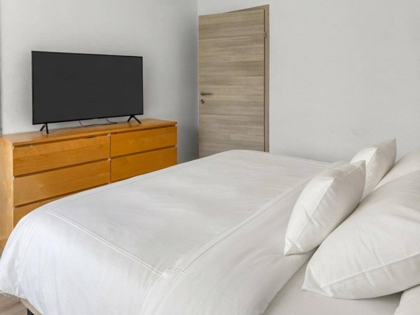 Schlafzimmer mit großem Fernseher auf einer Holzkommode und heller Holztür.