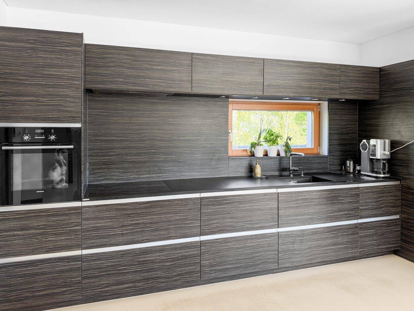 Moderne Einbauküche mit dunklen Holzfronten, integriertem Backofen und Fensterblick ins Grüne.