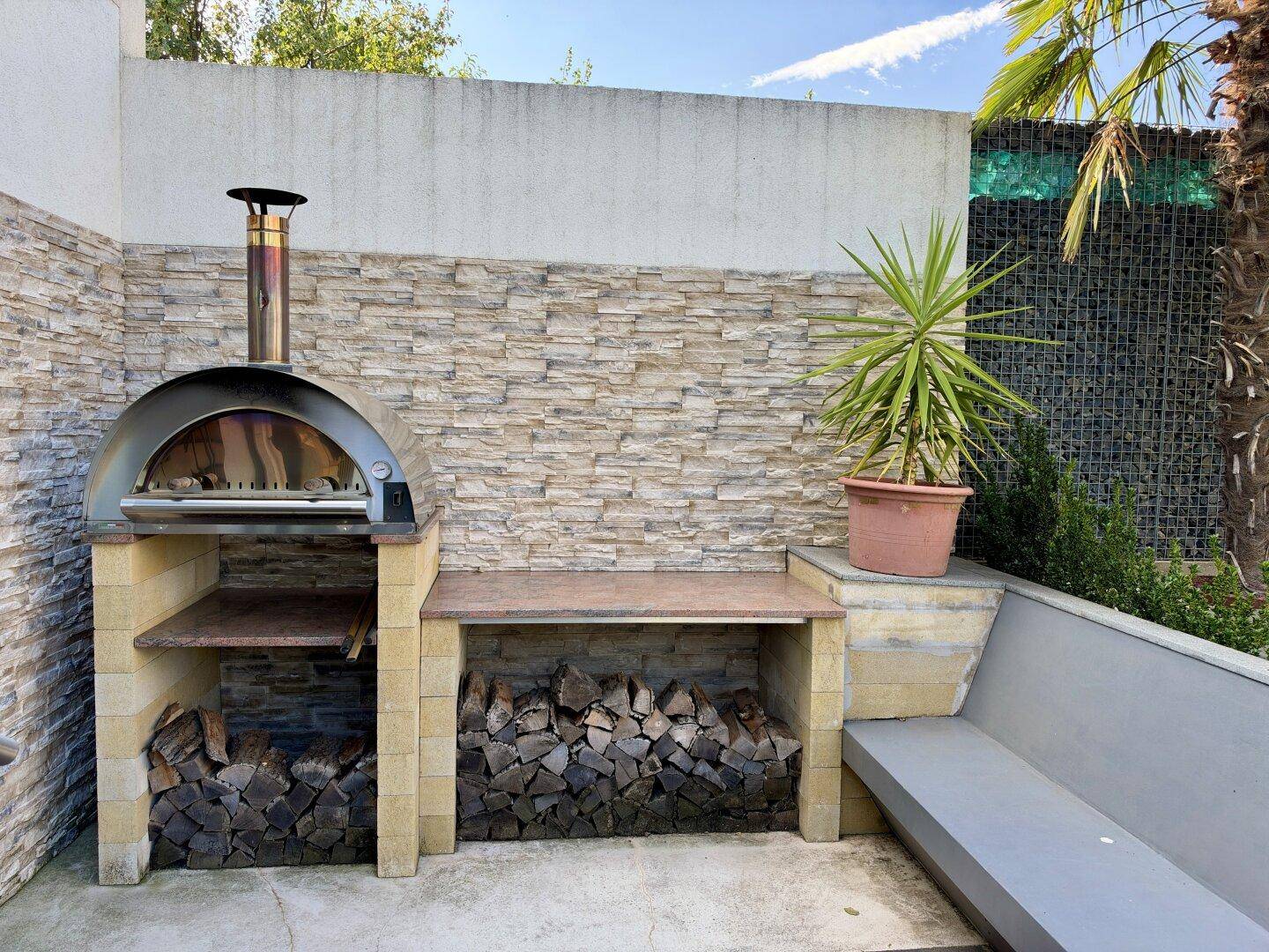 Gemütlicher Grillbereich mit Pizzaofen, Holzstapel und Sitzgelegenheit im Freien.
