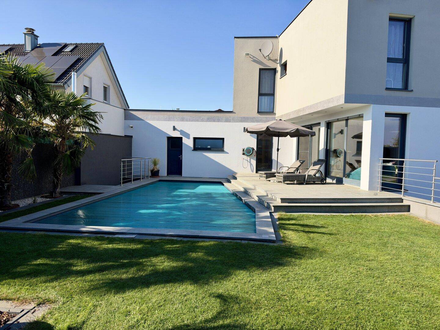 Weitläufiger Gartenbereich mit modernem Haus, Pool und Terrasse unter blauem Himmel.