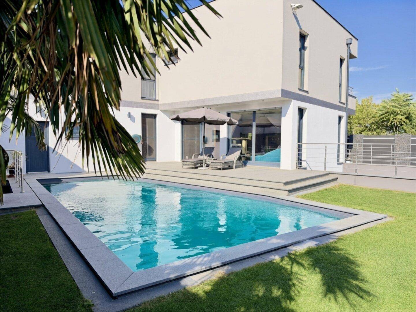 Rückansicht des modernen Hauses mit großem Swimmingpool, Rasenfläche und sonniger Terrasse.
