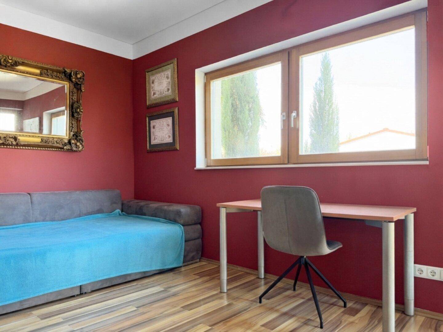 Farbenfrohes Zimmer mit roter Wand, Schlafsofa und Schreibtisch, ideal als Büro oder Gästezimmer.