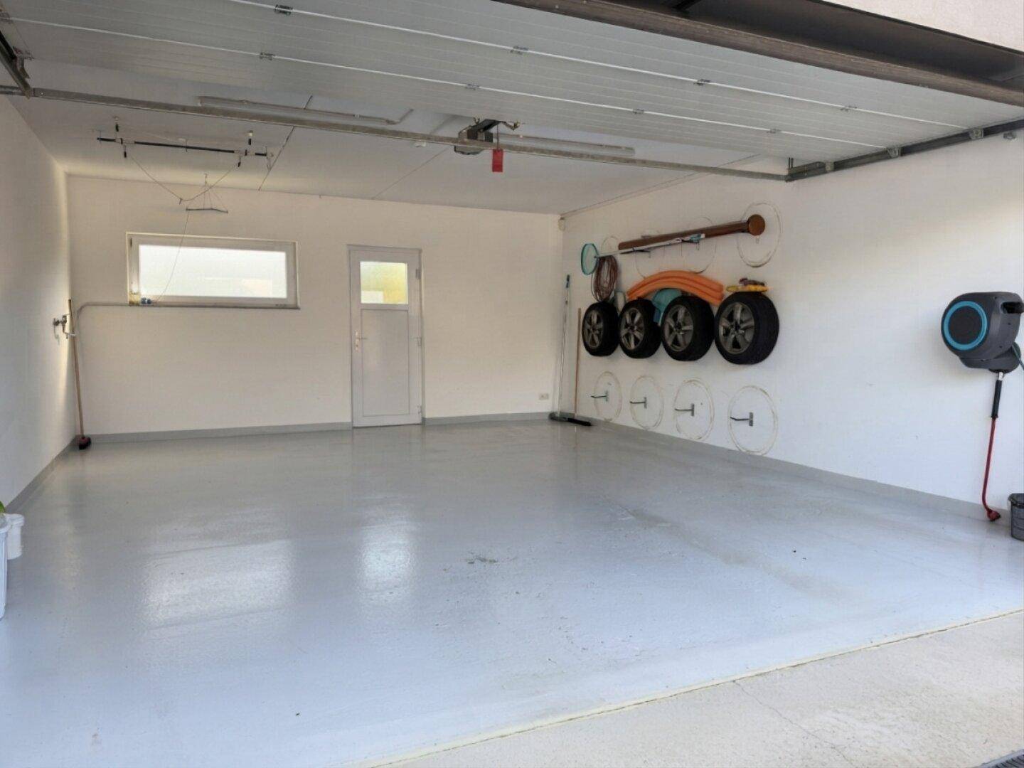 Leere Garage mit hellem Boden, Fenster und einer Tür zum Garten.