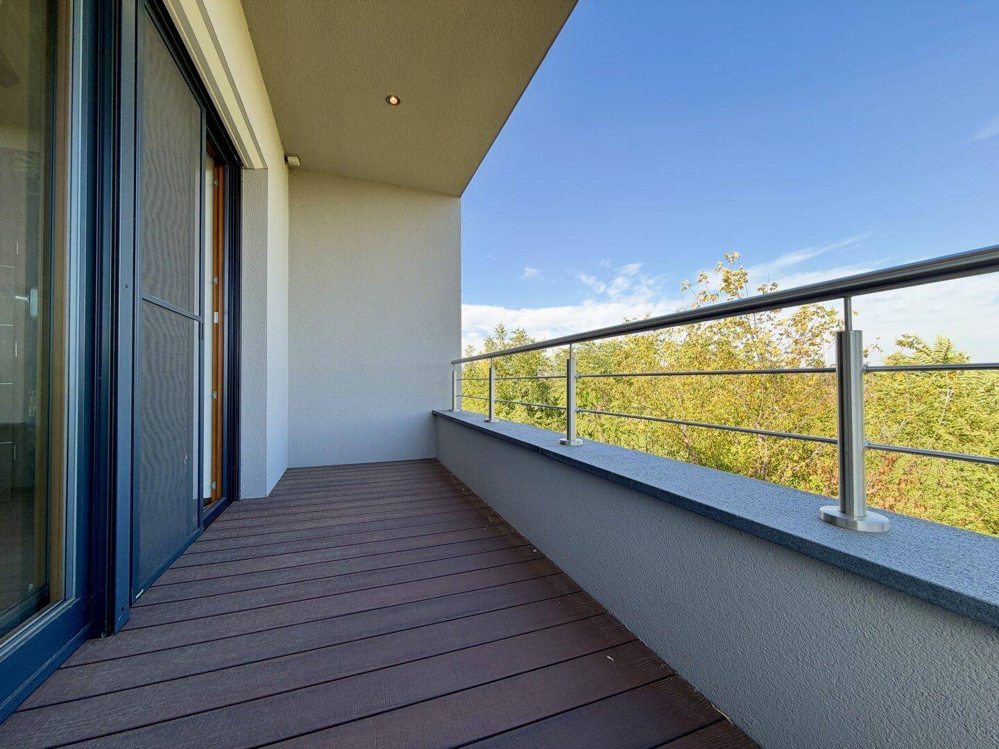 Geräumiger Balkon mit Holzboden und Metallgeländer, umgeben von grünen Bäumen.