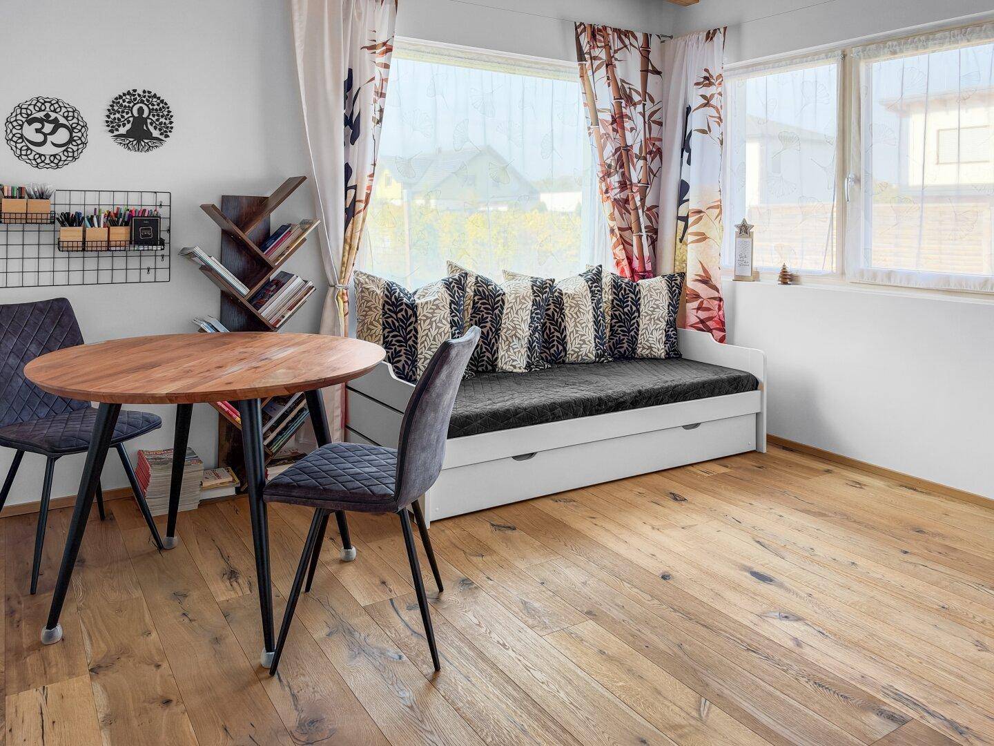 Helles Arbeitszimmer mit Holzboden, kleinem Tisch und Tagesbett am Fenster.