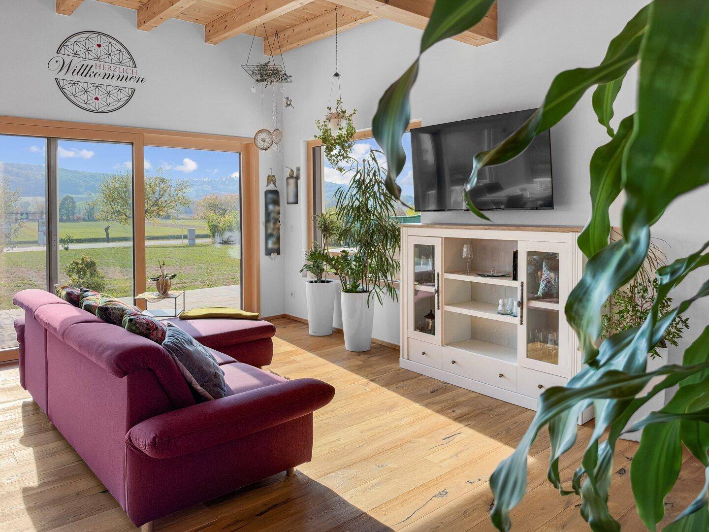 Lichtdurchflutetes Wohnzimmer mit Sofa, Fernseher und großen Fensterfronten, die einen weiten Ausblick bieten.