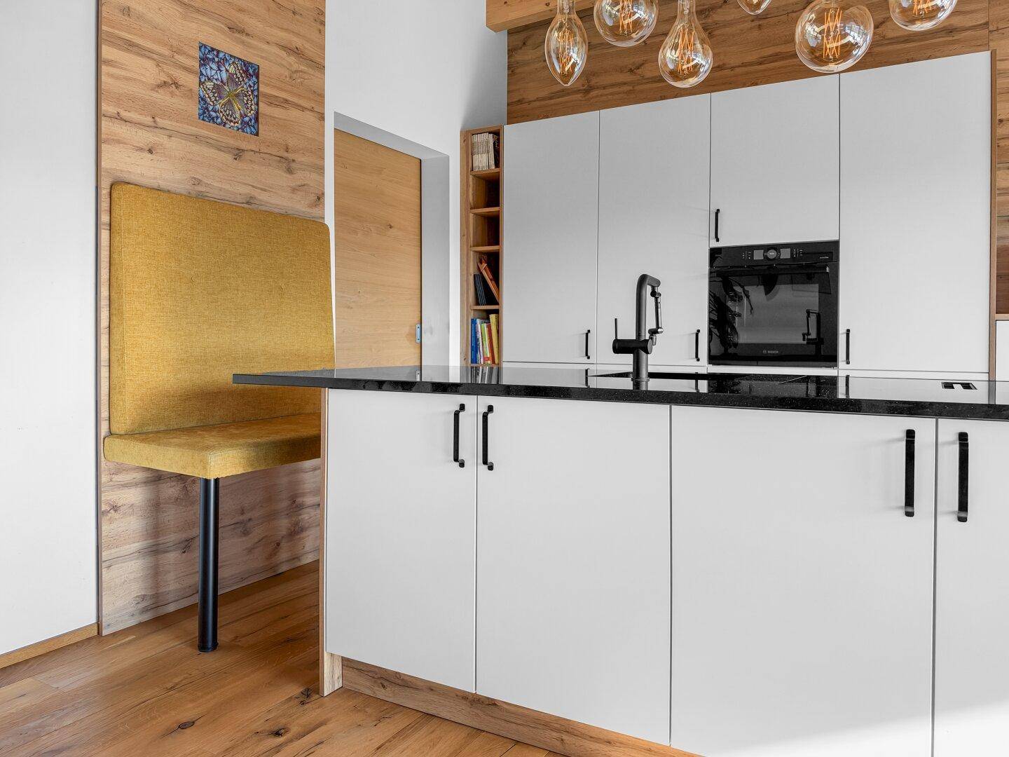 Moderne Kücheninsel mit schwarzer Arbeitsplatte, weißen Fronten und integriertem Sitzbereich vor Holzwand.