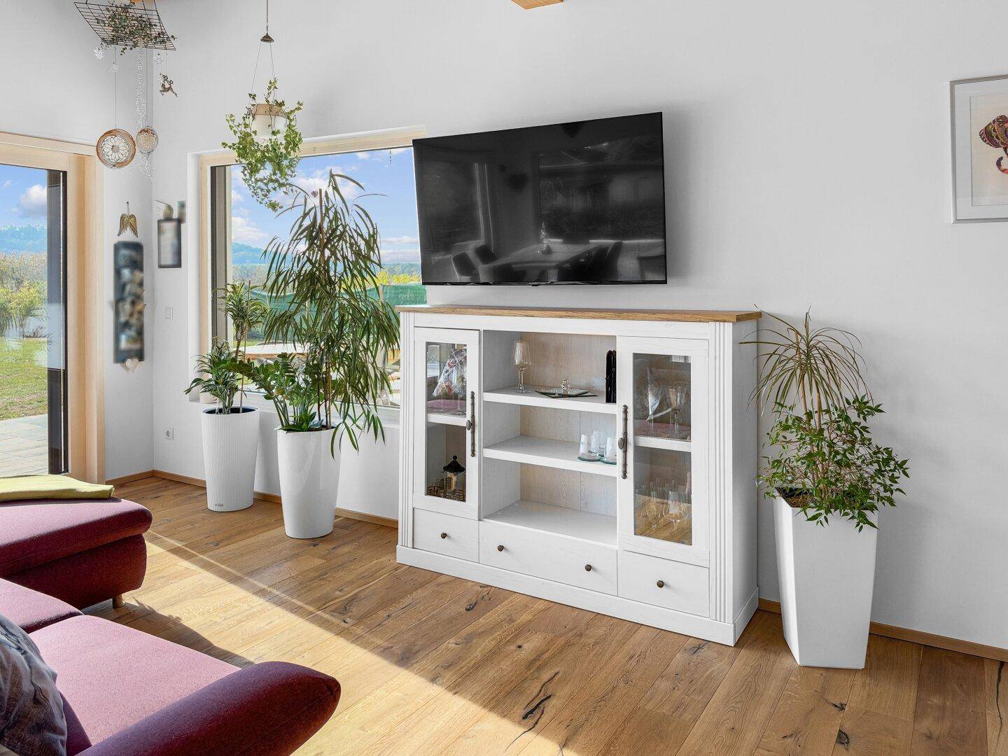 Wohnzimmer mit Holzboden, weißem Sideboard und großen Fenstern mit Blick ins Grüne.