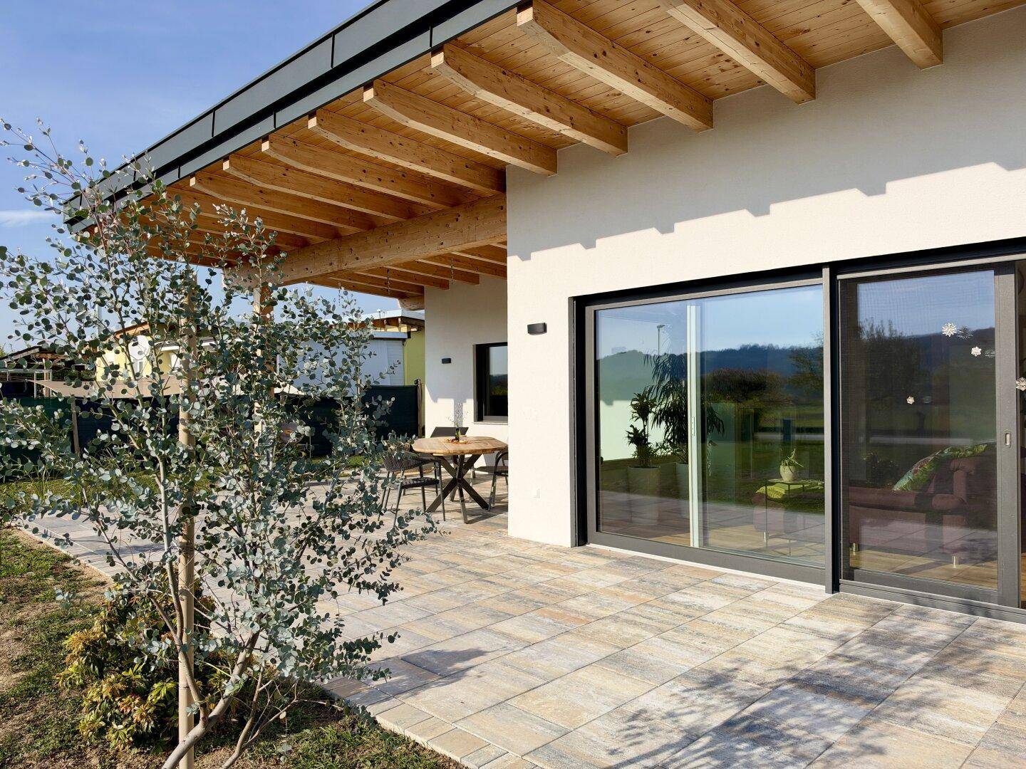 Helle Terrasse mit Holzbalkenüberdachung, großen Schiebetüren und Blick ins Grüne.