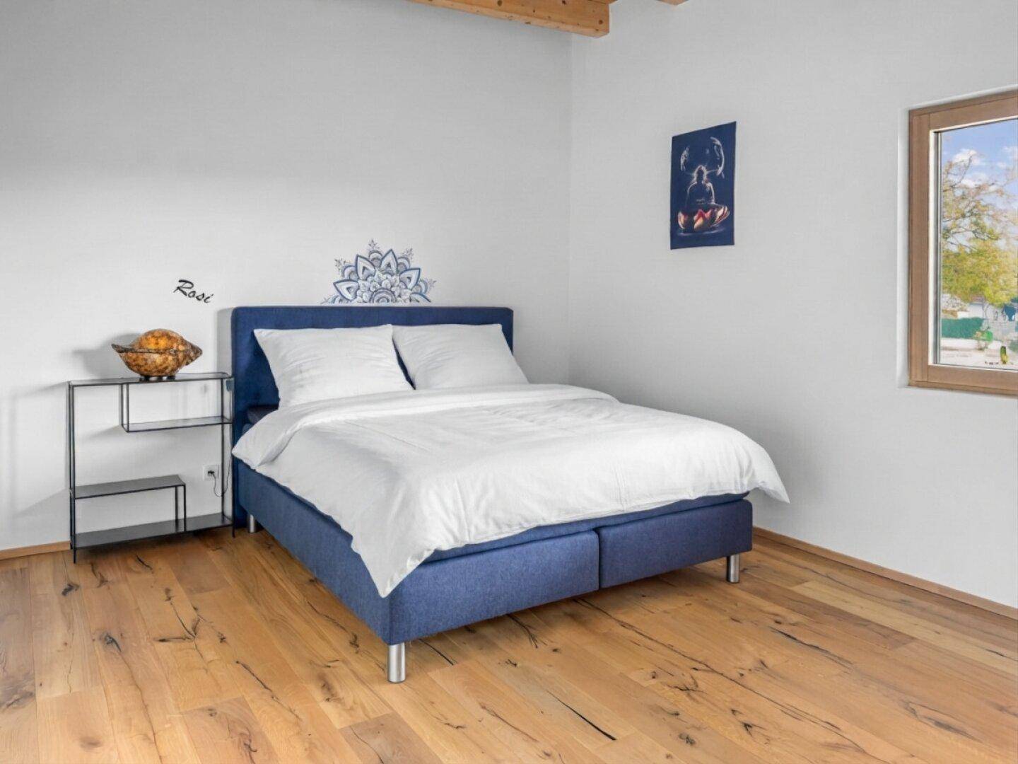 Schlafzimmer mit blauem Boxspringbett, Holzboden und Fenster mit Blick nach draußen.