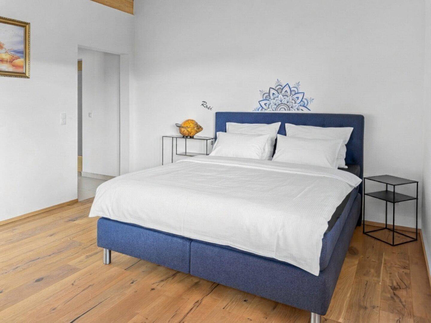 Helles Schlafzimmer mit blauem Bett, Holzboden und minimalistischer Einrichtung.