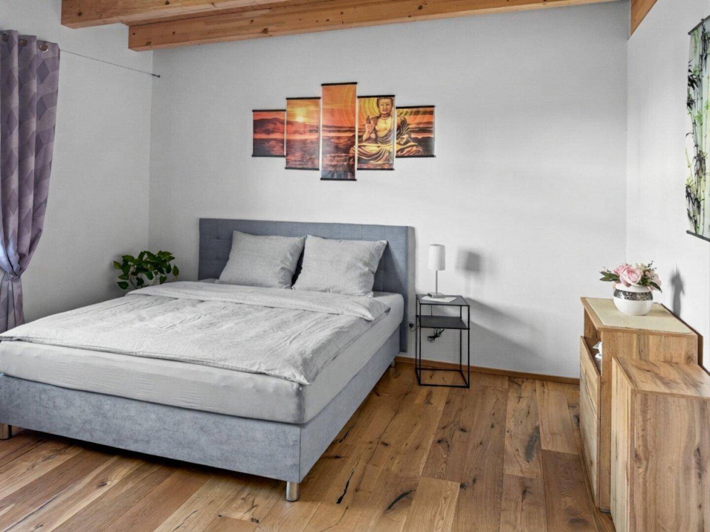 Schlafzimmer mit grauem Boxspringbett, Holzboden und dekorativer Wandkunst.