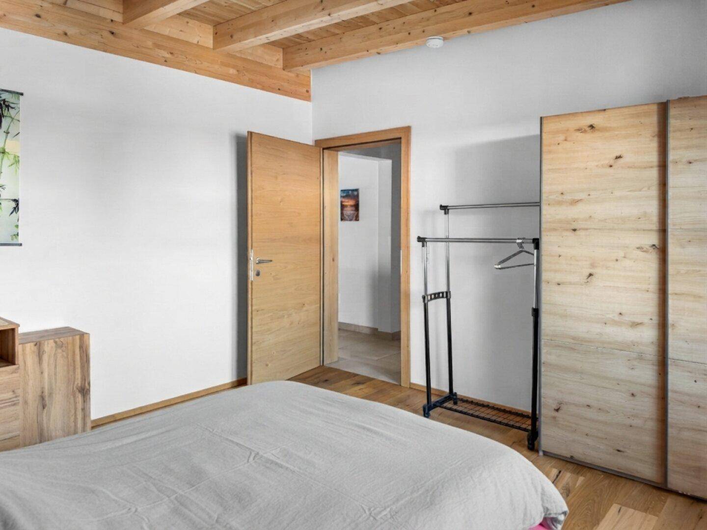 Schlafzimmer mit grauem Bett, Holzboden und offenem Kleiderständer.