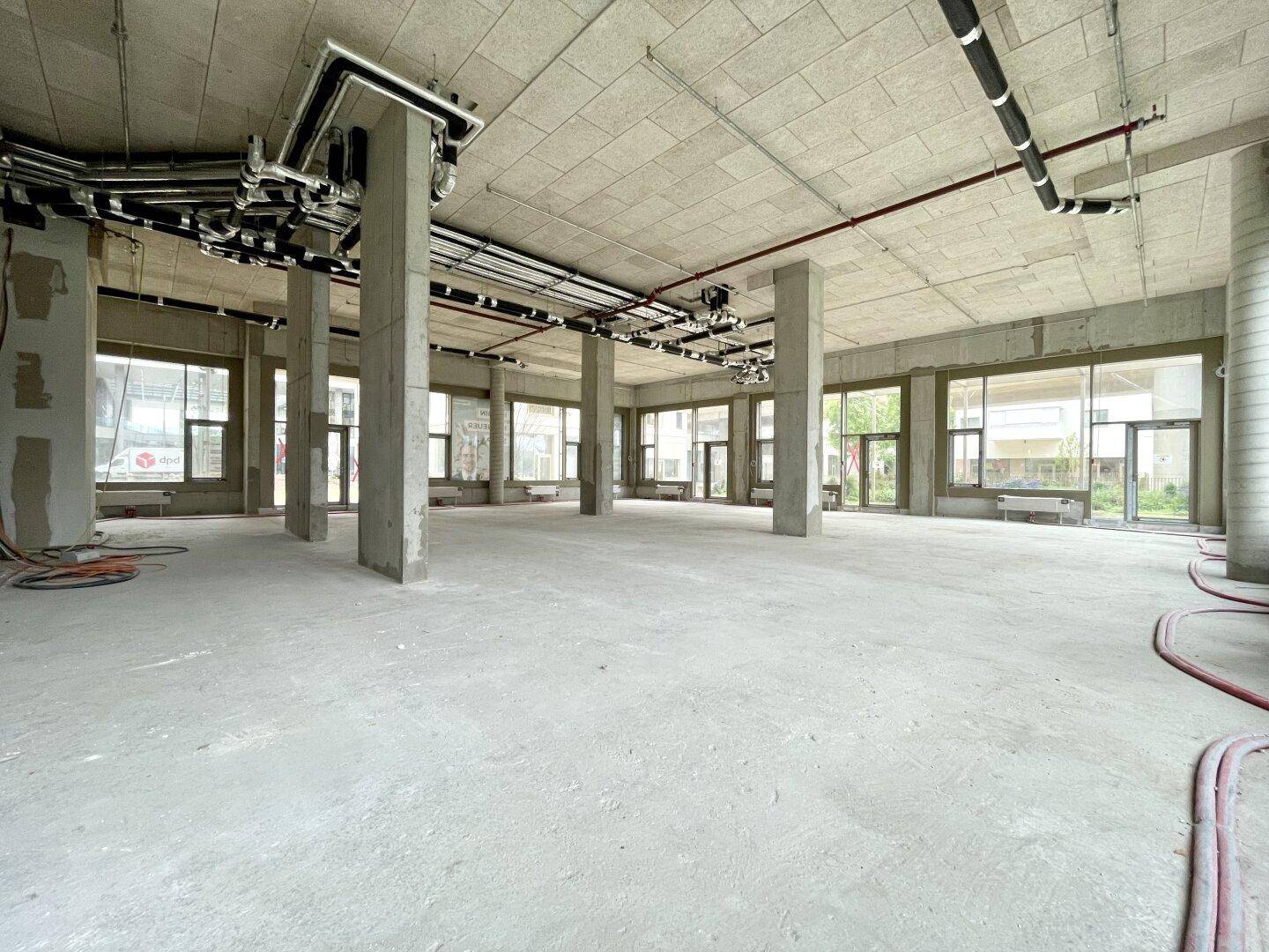 Großer, unfertiger Gewerberaum mit Betonboden, sichtbaren Installationen und vielen Fenstern.